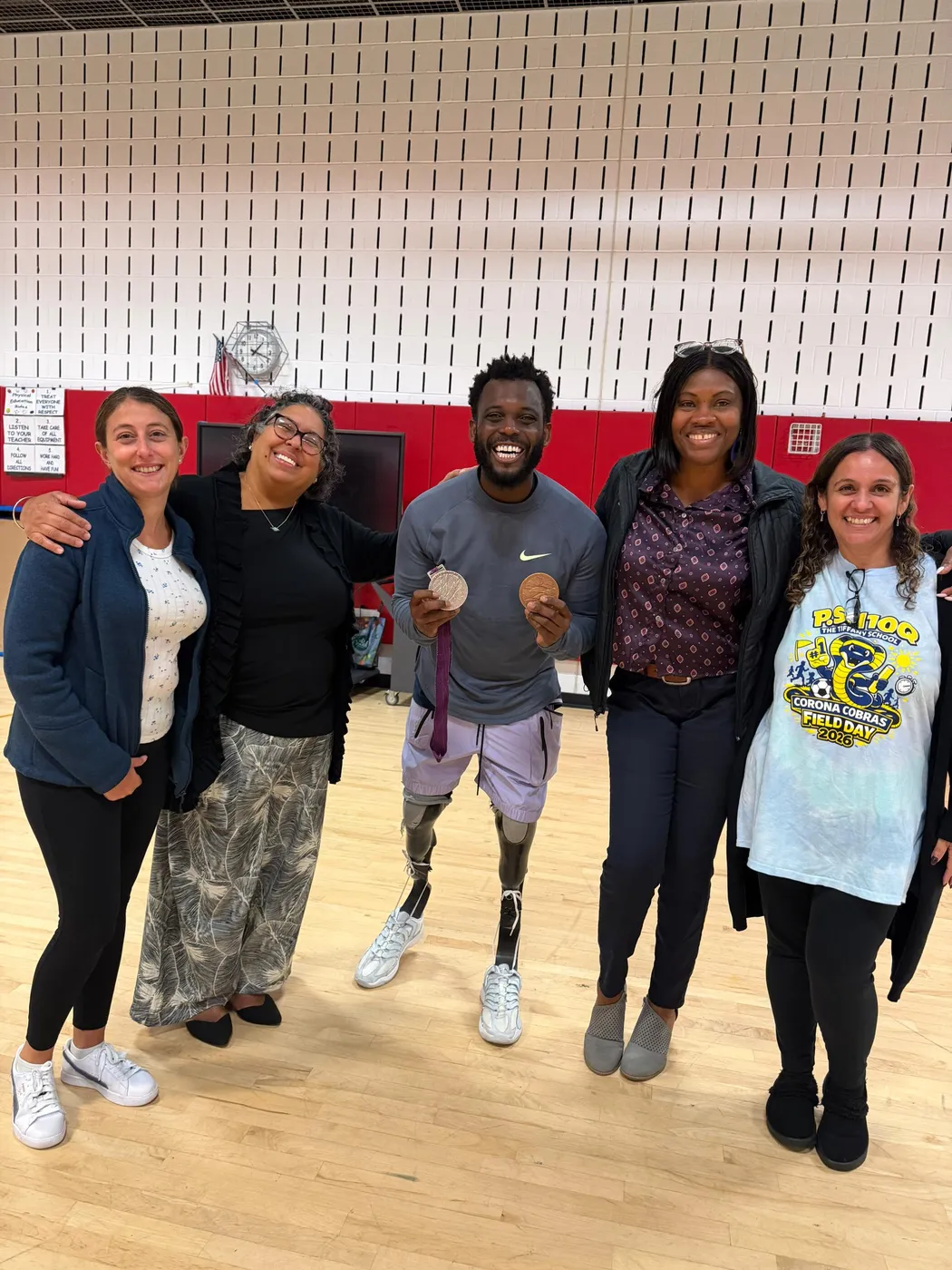 Paralympic sprinter Blake Leeper holding two medals in the P.S. 110Q gym, smiling with four members of the school's staff and administration